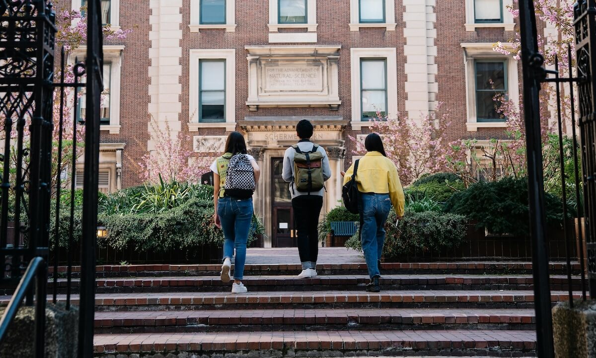 Three people with book bags walk up steps to a building together.